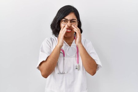 Téléchargez les photos : Young hispanic doctor woman wearing stethoscope over isolated background smelling something stinky and disgusting, intolerable smell, holding breath with fingers on nose. bad smell - en image libre de droit