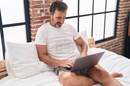 Foto de Hombre de mediana edad usando portátil sentado en la cama en el dormitorio - Imagen libre de derechos