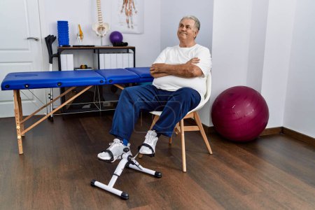Photo for Senior caucasian man at physiotherapy clinic using pedal exerciser smiling looking to the side and staring away thinking. - Royalty Free Image