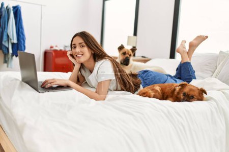 Téléchargez les photos : Jeune femme hispanique utilisant un ordinateur portable couché sur le lit avec des chiens dans la chambre - en image libre de droit