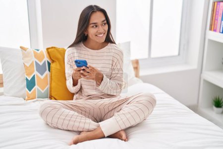Foto de Mujer hispana hermosa joven usando teléfono inteligente sentado en la cama en el dormitorio - Imagen libre de derechos