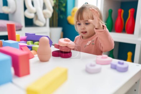 Foto de Adorable chica rubia jugando con bloques de construcción sentado en la mesa en el jardín de infantes - Imagen libre de derechos