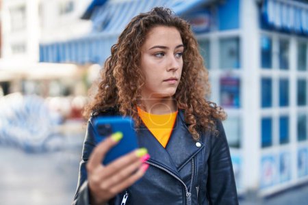 Foto de Mujer hispana hermosa joven usando teléfono inteligente con expresión relajada en la calle - Imagen libre de derechos