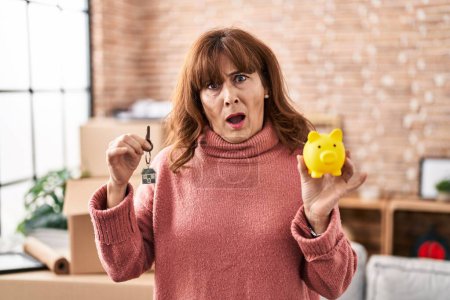 Photo for Middle age hispanic woman holding piggy bank and house keys in shock face, looking skeptical and sarcastic, surprised with open mouth - Royalty Free Image