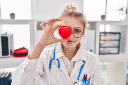 Photo for Young blonde woman doctor holding heart over eye at clinic - Royalty Free Image