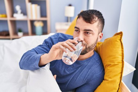 Young hispanic man drinking water lying on bed at bedroom