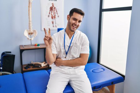 Photo for Young hispanic man with beard working at pain recovery clinic smiling with happy face winking at the camera doing victory sign. number two. - Royalty Free Image