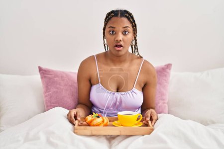 Téléchargez les photos : African american woman with braids holding tray with breakfast food in the bed scared and amazed with open mouth for surprise, disbelief face - en image libre de droit