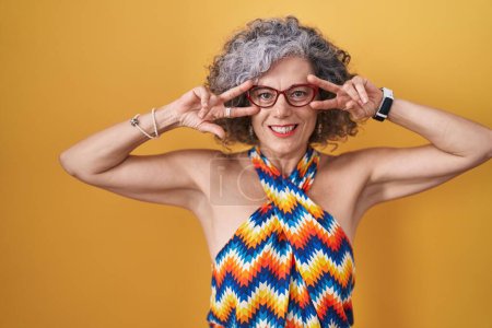 Téléchargez les photos : Femme d'âge moyen avec les cheveux gris debout sur fond jaune faisant symbole de paix avec les doigts sur le visage, souriant joyeuse montrant la victoire - en image libre de droit