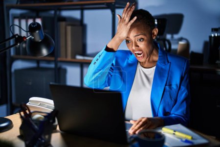 Téléchargez les photos : Beautiful african american woman working at the office at night surprised with hand on head for mistake, remember error. forgot, bad memory concept. - en image libre de droit