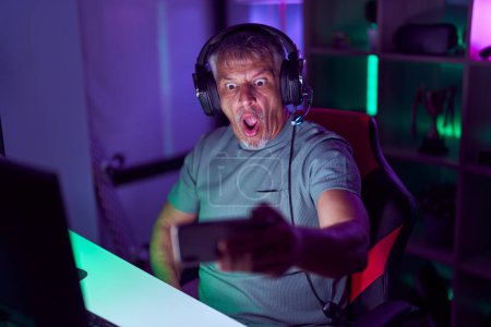 Foto de Hombre hispano con pelo gris jugando videojuegos con smartphone asustado y sorprendido con la boca abierta para sorpresa, cara de incredulidad - Imagen libre de derechos