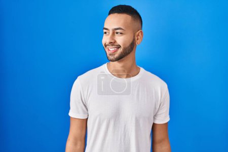 Téléchargez les photos : Young hispanic man standing over blue background looking away to side with smile on face, natural expression. laughing confident. - en image libre de droit