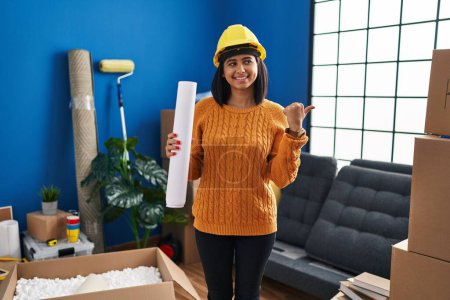 Photo for Young hispanic woman standing on new home with blueprints pointing thumb up to the side smiling happy with open mouth - Royalty Free Image