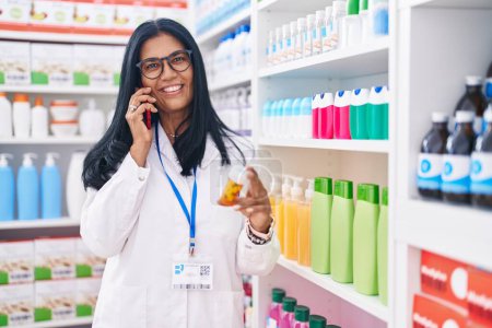Téléchargez les photos : Moyen Age hispanique femme pharmacien tenant bouteille pilules parler sur smartphone à la pharmacie - en image libre de droit