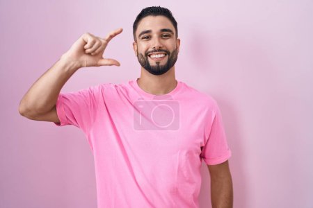 Téléchargez les photos : Hispanic young man standing over pink background smiling and confident gesturing with hand doing small size sign with fingers looking and the camera. measure concept. - en image libre de droit