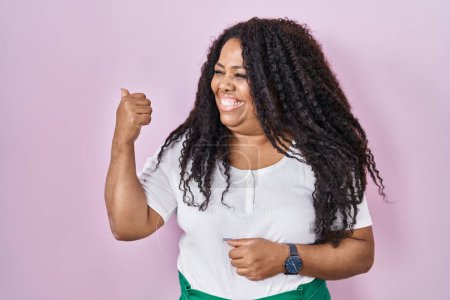 Téléchargez les photos : Plus size hispanic woman standing over pink background smiling with happy face looking and pointing to the side with thumb up. - en image libre de droit