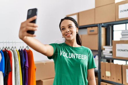 Photo for Young african american woman wearing volunteer uniform make selfie by the smartphone at charity center - Royalty Free Image