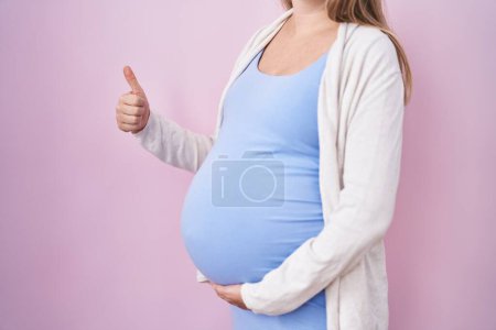 Photo for Young pregnant woman expecting a baby, touching pregnant belly smiling happy and positive, thumb up doing excellent and approval sign - Royalty Free Image