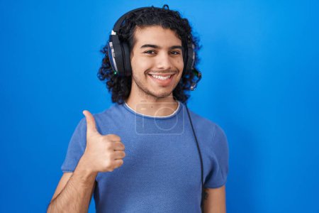 Photo for Hispanic man with curly hair listening to music using headphones doing happy thumbs up gesture with hand. approving expression looking at the camera showing success. - Royalty Free Image