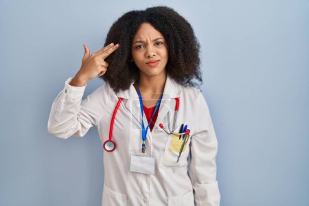 Foto de Joven mujer afroamericana vistiendo uniforme médico y estetoscopio disparando y matándose señalando la mano y los dedos a la cabeza como arma, gesto suicida. - Imagen libre de derechos