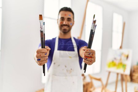 Photo for Young hispanic man smiling confident holding paintbrushes at art studio - Royalty Free Image