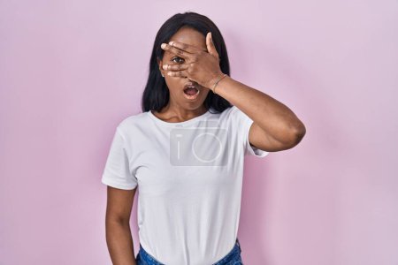 Téléchargez les photos : Jeune femme africaine vêtue d'un t-shirt blanc décontracté jetant un coup d'œil sur le visage et les yeux avec la main, regardant à travers les doigts avec une expression embarrassée. - en image libre de droit