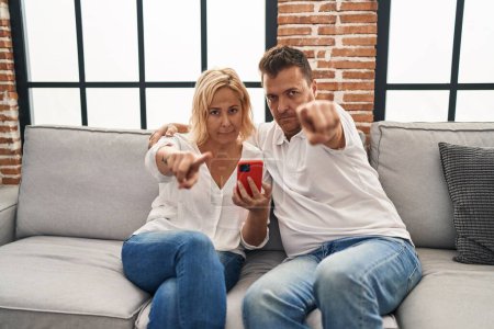 Foto de Hombre y mujer de mediana edad usando un teléfono inteligente sentado en el sofá señalando con el dedo a la cámara y a usted, gesto de confianza que se ve serio - Imagen libre de derechos