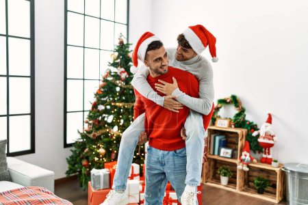 Téléchargez les photos : Deux hommes hispaniques couple tenant sur le dos debout près de l'arbre de Noël à la maison - en image libre de droit