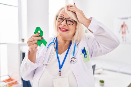 Foto de Middle age caucasian woman holding support green ribbon stressed and frustrated with hand on head, surprised and angry face - Imagen libre de derechos
