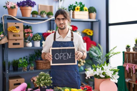 Photo for Hispanic man with long hair working at florist holding open sign clueless and confused expression. doubt concept. - Royalty Free Image
