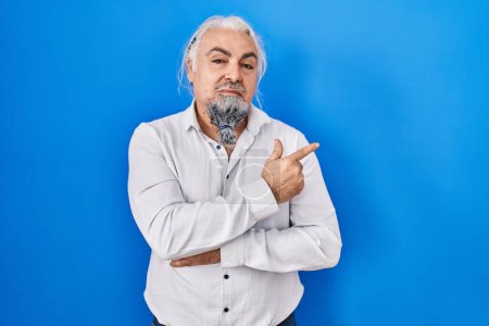Foto de Middle age man with grey hair standing over blue background pointing with hand finger to the side showing advertisement, serious and calm face - Imagen libre de derechos