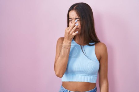Téléchargez les photos : Jeune femme brune debout sur fond rose sentant quelque chose de puant et dégoûtant, odeur intolérable, retenant la respiration avec les doigts sur le nez. mauvaise odeur - en image libre de droit
