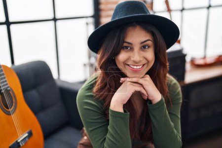 Téléchargez les photos : Jeune musicienne hispanique souriante confiante assise sur un canapé au studio de musique - en image libre de droit