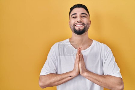 Téléchargez les photos : Young handsome man wearing casual t shirt over yellow background praying with hands together asking for forgiveness smiling confident. - en image libre de droit