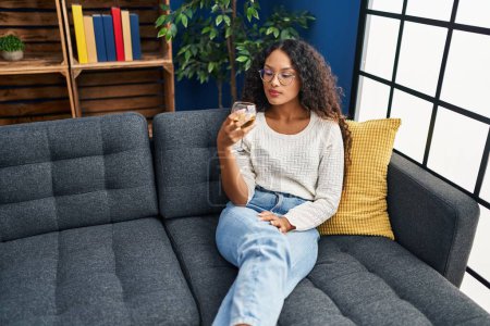 Téléchargez les photos : Jeune femme latine buvant du champagne assise sur un canapé à la maison - en image libre de droit
