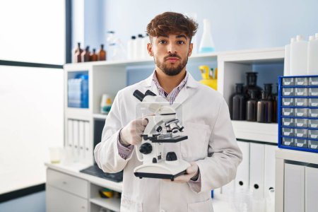 Téléchargez les photos : Homme arabe avec barbe travaillant au laboratoire scientifique tenant un microscope détendu avec une expression sérieuse sur le visage. simple et naturel regardant la caméra. - en image libre de droit