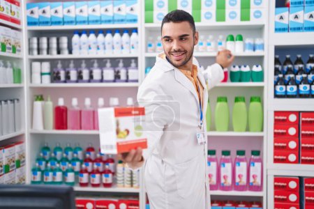 Foto de Joven farmacéutico hispano sonriendo confiado sosteniendo paquete de vitaminas en la farmacia - Imagen libre de derechos