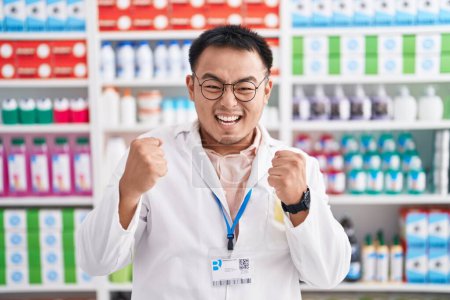 Téléchargez les photos : Chinese young man working at pharmacy drugstore celebrating surprised and amazed for success with arms raised and eyes closed. winner concept. - en image libre de droit