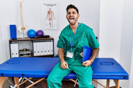 Photo for Young physiotherapist man working at pain recovery clinic sticking tongue out happy with funny expression. emotion concept. - Royalty Free Image