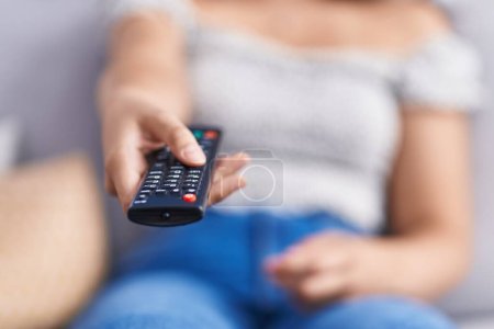 Photo for Young beautiful hispanic woman using tv remote control sitting on sofa at home - Royalty Free Image