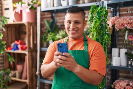 Téléchargez les photos : Jeune homme latin fleuriste souriant confiant en utilisant un smartphone à la boutique de fleurs - en image libre de droit