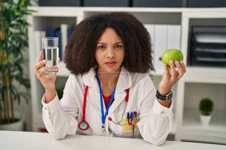Foto de Young african american dietitian woman holding fresh apple and water clueless and confused expression. doubt concept. - Imagen libre de derechos