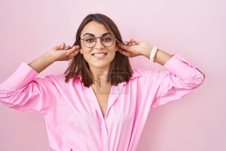 Foto de Mujer hispana joven con gafas de pie sobre fondo rosa sonriendo tirando de las orejas con los dedos, gesto divertido. problema de audición - Imagen libre de derechos