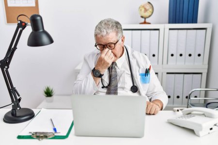 Téléchargez les photos : Senior caucasian man wearing doctor uniform and stethoscope at the clinic tired rubbing nose and eyes feeling fatigue and headache. stress and frustration concept. - en image libre de droit