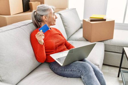 Téléchargez les photos : Middle age grey-haired woman smiling confident using laptop and credit card at new home - en image libre de droit