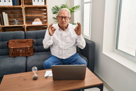 Téléchargez les photos : Senior therapist with grey hair doing online session at consultation office relax and smiling with eyes closed doing meditation gesture with fingers. yoga concept. - en image libre de droit