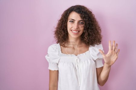 Photo for Hispanic woman with curly hair standing over pink background showing and pointing up with fingers number five while smiling confident and happy. - Royalty Free Image
