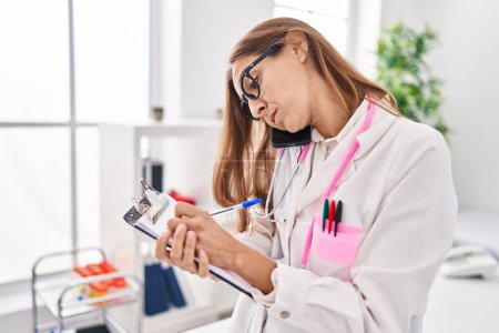 Foto de Young woman wearing doctor uniform talking on the smartphone write on clipboard at clinic - Imagen libre de derechos