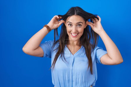Téléchargez les photos : Jeune femme brune debout sur fond bleu souriant tirant les oreilles avec les doigts, geste drôle. problème d'audition - en image libre de droit