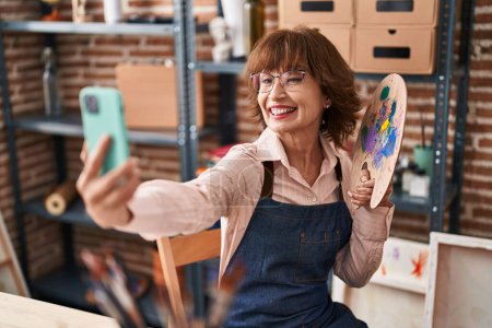 Téléchargez les photos : Femme d'âge moyen artiste souriant confiant faire selfie par smartphone au studio d'art - en image libre de droit